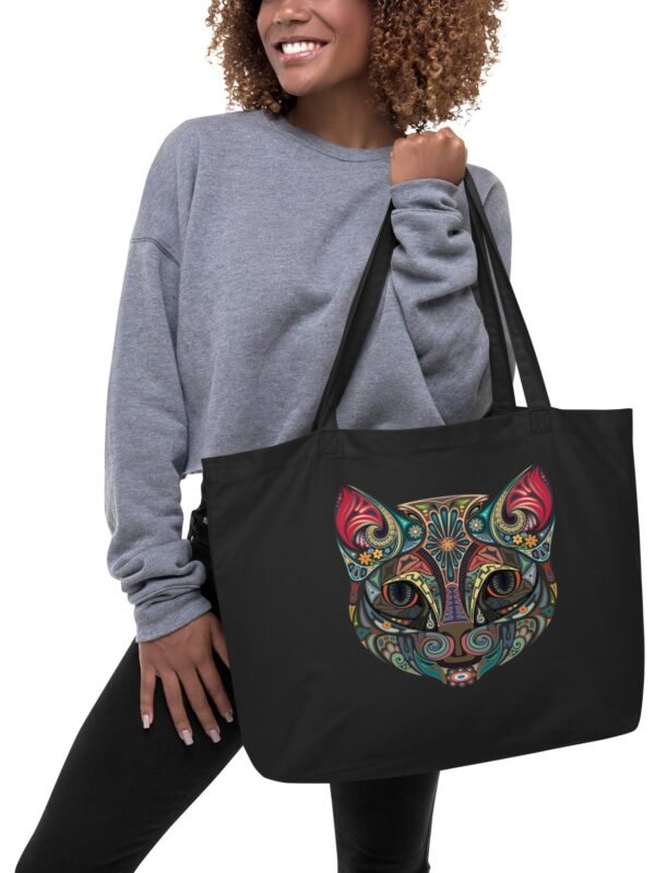 A person holding a black organic tote bag adorned with a colorful tribal cat design.